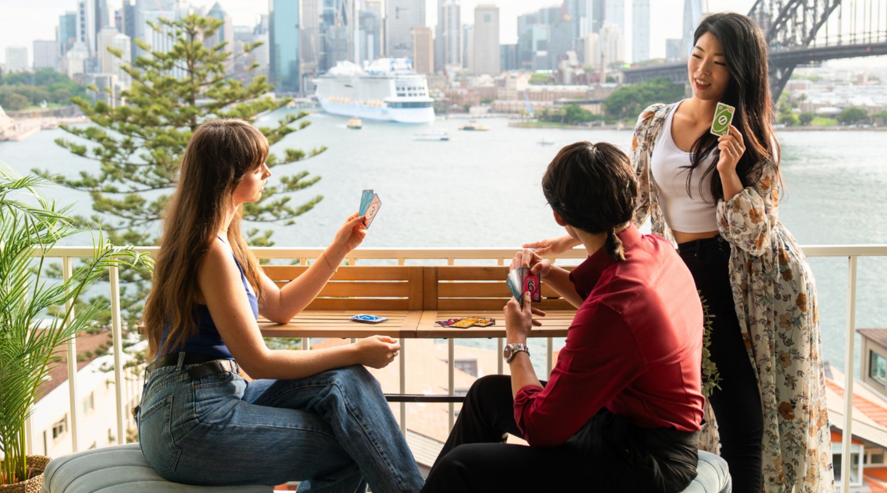 Three people playing cards on a views balcony bar table with a cityscape and harbor view.